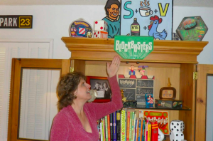 Cathi Ryan, Steve's wife, poses with some of his game show memorabilia, including the Blockbusters home game, logo from the front of Bill Cullen's podium, and the preserved slate from the final taping